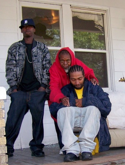 The morning of Aiyana's death, Charles Jones is comforted by his aunt JoAnn Robinson, now deceased, and her son Mark Robinson. Mark warned the raid team there were children in the house before they entered; toys were strewn on the front yard. Behind Mark's head is window shattered by stun grenade; couch where Aiyana died is next to Charles.