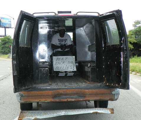 Baxter Jones, in battered police truck, proclaims "Beat Back the Bullies."